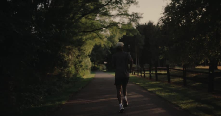 Male jogger runs along a path in the park passing a grove and a fenced lawn
