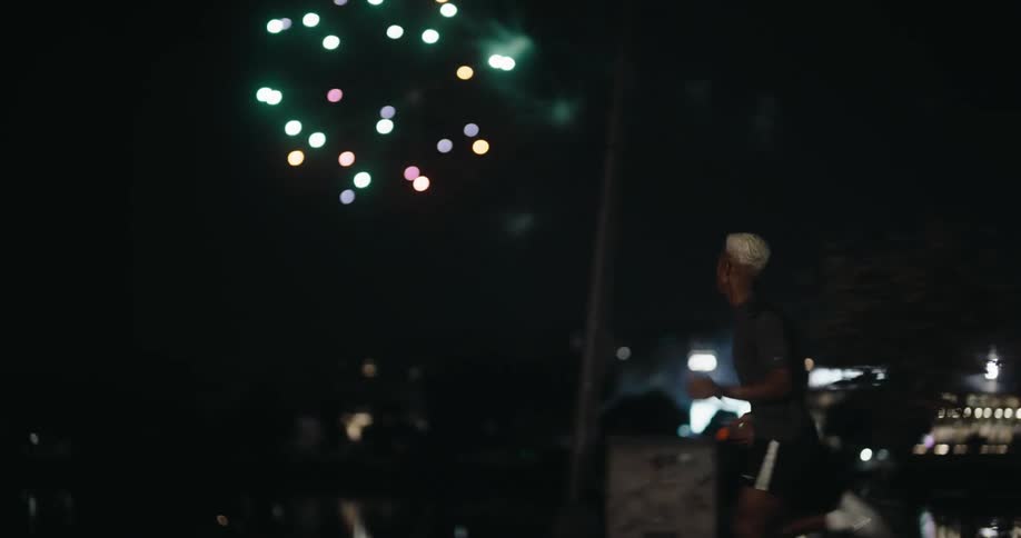 A young man jogs at night with fireworks exploding in the distance