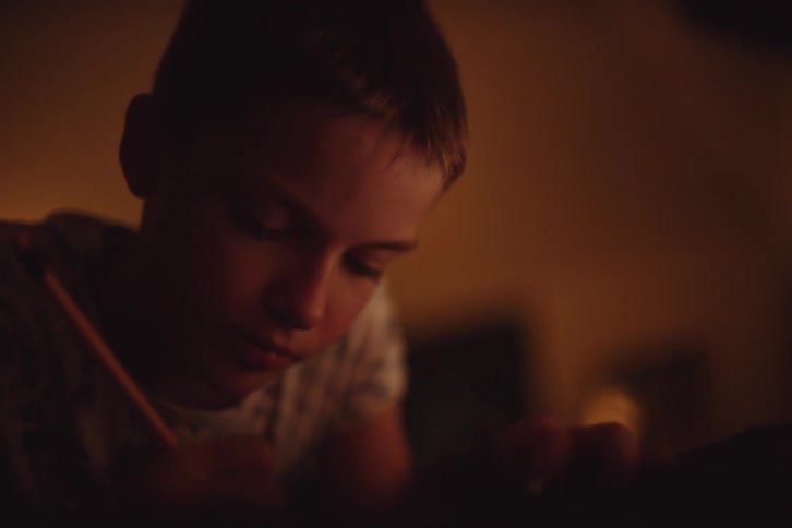 A young boy sits in a bedroom while writing something with a pencil