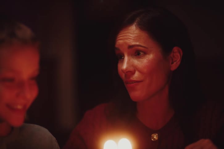 A mother carrying a lit birthday cake enters a darkened room singing a happy birthday song to her son