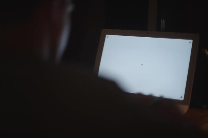 A person sits at a laptop in a dark room with the screen displaying a blank white background