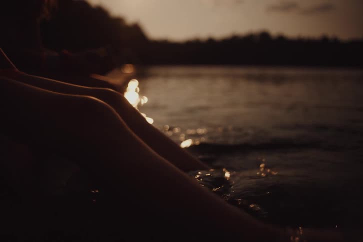 A boy and a girl sit by the waters edge their legs dangling in the water as the sun sets behind them