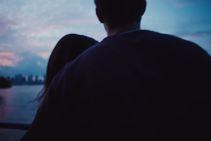 A woman looks up happily at her boyfriend by the sea as the sun sets over the city skyline