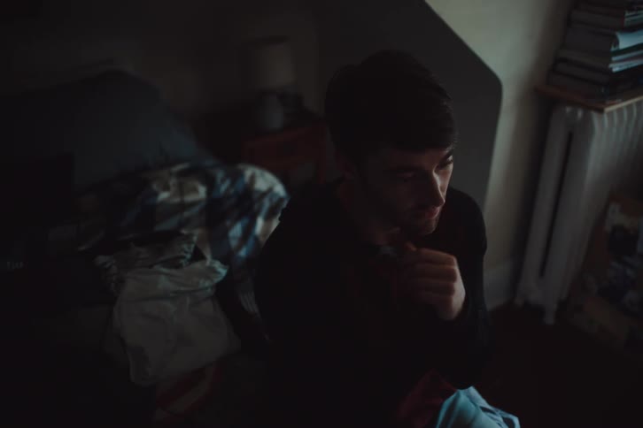 A man sits in a dimly lit bedroom resting his face on his hand with a thoughtful expression surrounded by scattered clothing and subtle shadows that add to the somber mood of the scene