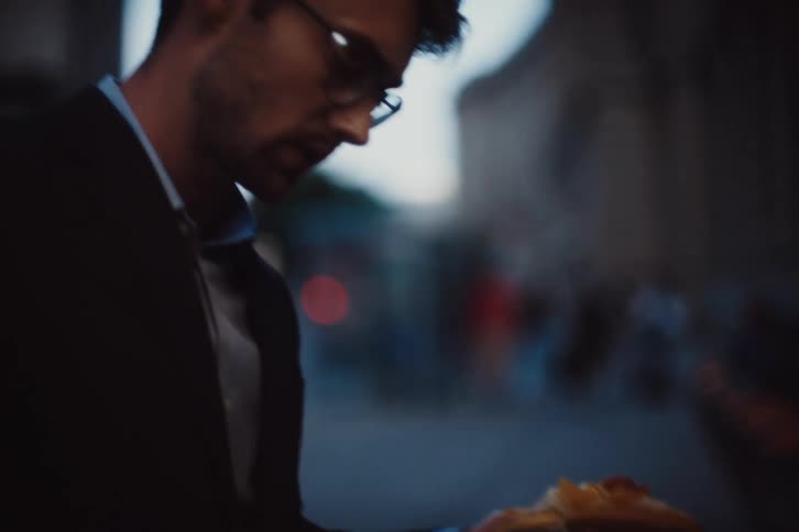 A man in a suit holding a hot dog talks to someone on a city street at dusk