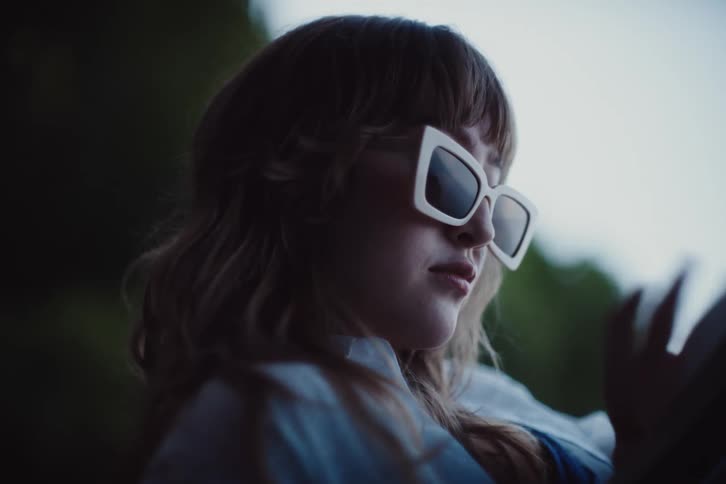 The woman with wavy hair wearing oversized white sunglasses reads a book set against a blurred outdoor background