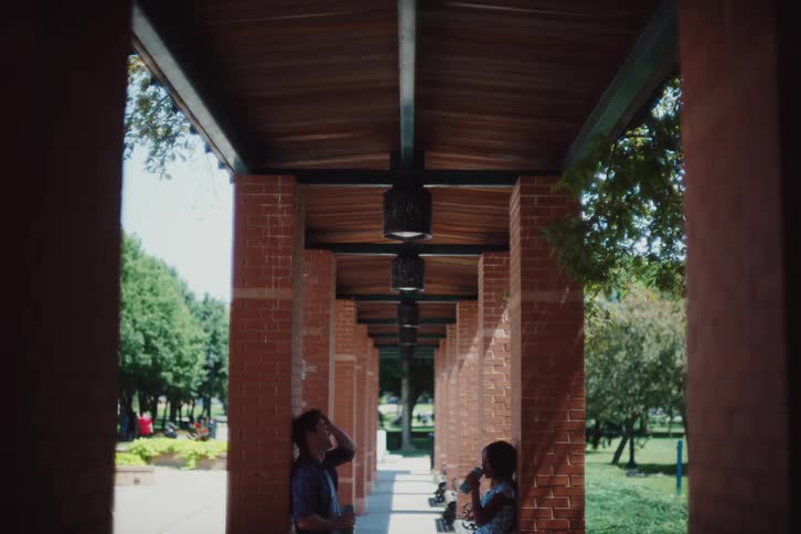 A shaded walkway with brick columns and a wooden ceiling where two individuals stand apart each leaning against a column and drinking beverages