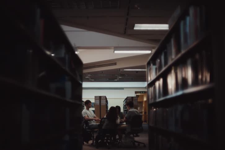 A group of people sitting at a table in a library seen from between two rows of bookshelves with overhead lighting and a quiet focused atmosphere