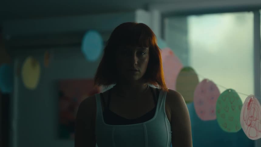 A woman with shoulderlength red hair standing in a dimly lit room wearing a white tank top with colorful decorations visible in the blurred background