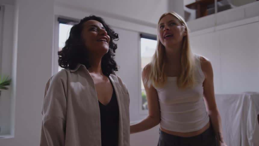 Two women one carrying a box and the other a potted plant placing them in a living room and both hugging each other