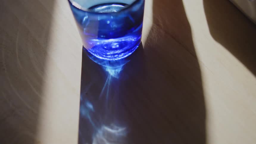 Hand placing a blue cup of water on a wooden surface with sunlight reflecting off it
