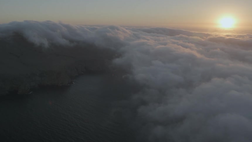 The sun sets above clouds covering water and hills