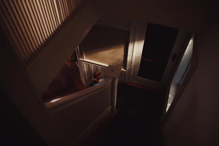 A child sits on a staircase in a dimly lit hallway opening a letter and reading it