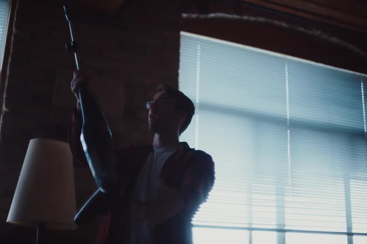 A man in a burgundy shirt and glasses is dancing with a vacuum cleaner in a dimly lit room with windows