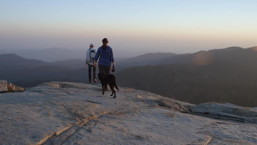 Two people walking on a rocky mountain top with their dog and coming to rest at the edge