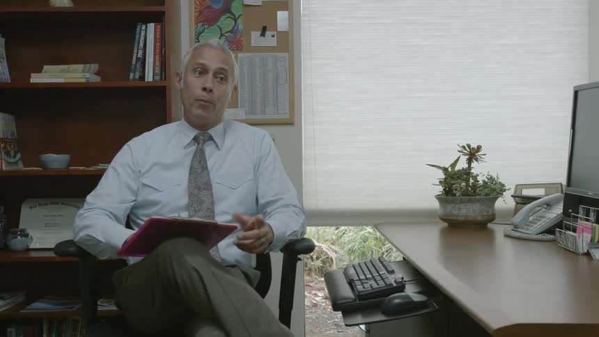 A man holding a clipboard is sitting near a desk and speaking at a woman