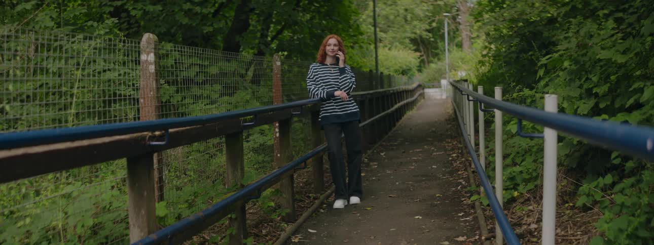 A woman in a striped shirt is leaning against the railing of a bridge in the middle of the forest
