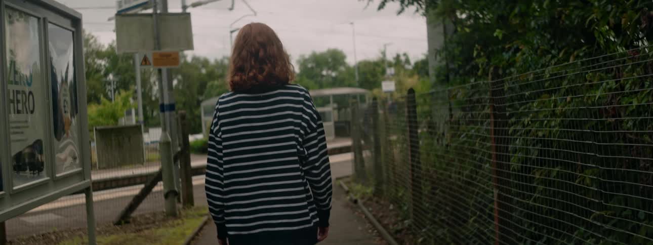 A redheaded female walks down a sidewalk that runs in between a fence and a display case for posters