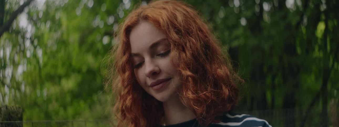 Redhead woman with happy facial expression looking down and surrounded by trees