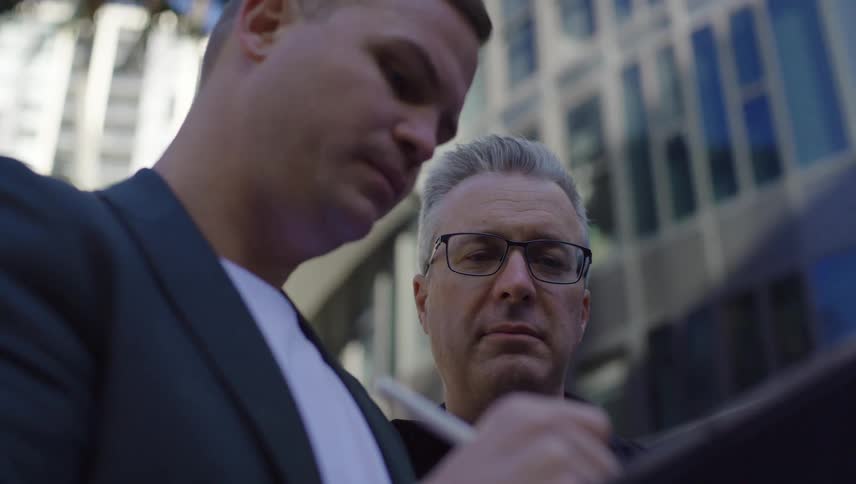 A man is writing something down in a notebook with a pen and he is wearing a suit and another man behind him is looking at the notebook intently