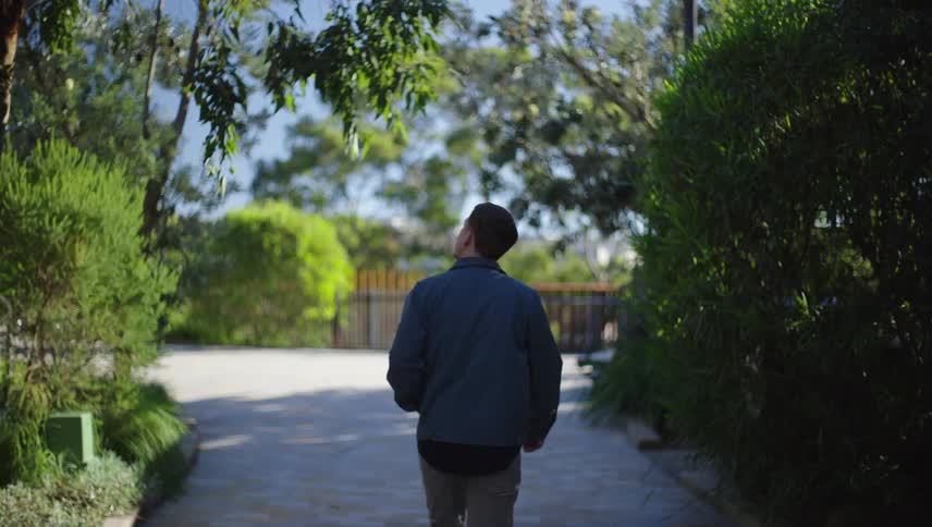 A man in a blue jacket is walking on a concrete path surrounded by greenery and some trees