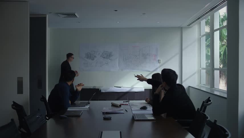 A person drawing on a paper on a wall in a meeting room and there are three people pointing to drawings and talking about
