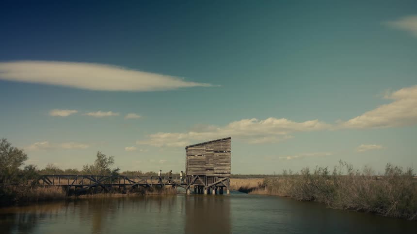 A wooden structure on stilts over a calm body of water with people walking across a walkway leading to it and a partly cloudy sky