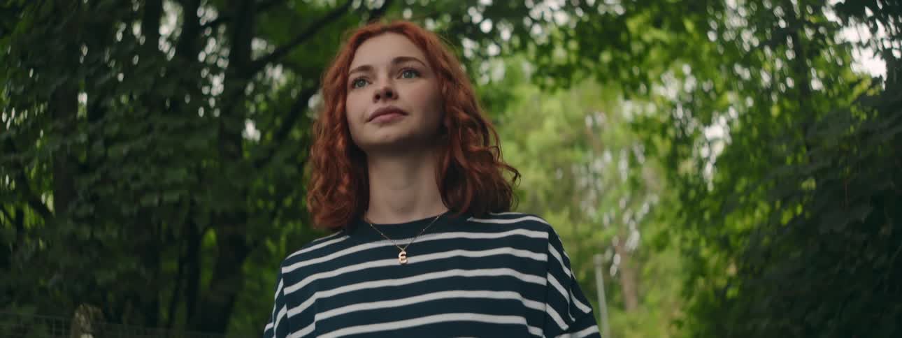 A teenage girl with red hair smiles slightly as she walks through a forest