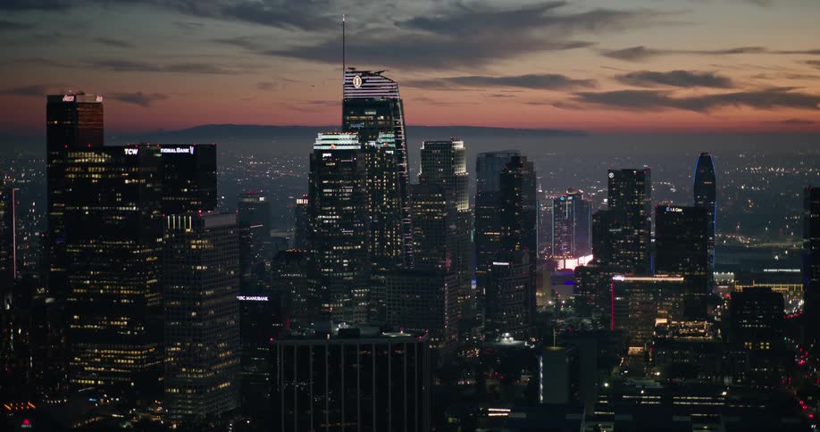 An aerial view of the los angeles city skyline at twilight with illuminated skyscrapers and a partly cloudy sky