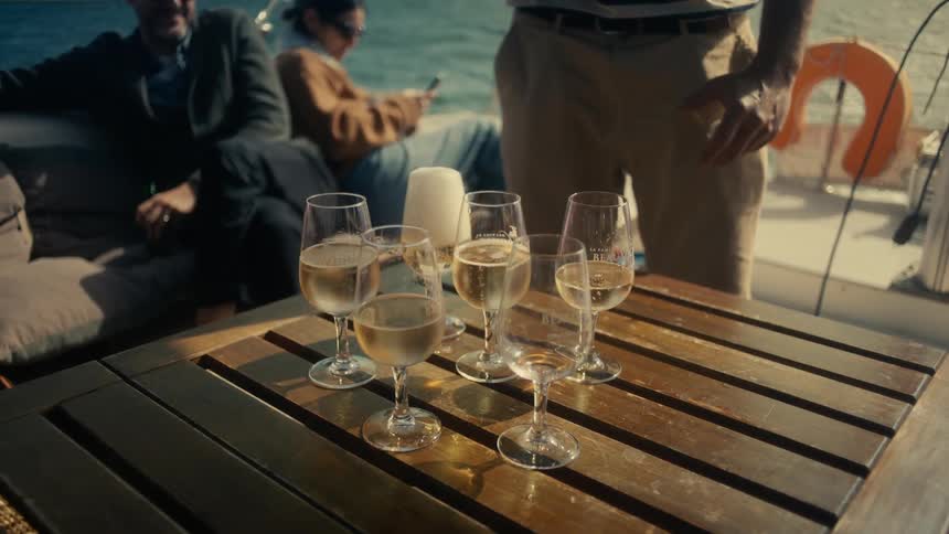 A wooden table on a boat with multiple tall glasses of champagne on it while a man pours white wine from a bottle and two other people sit waiting for the glasses
