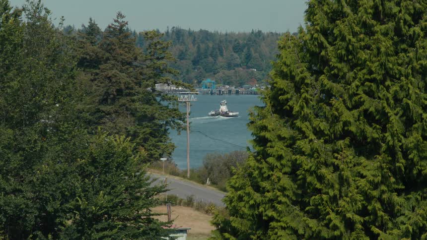 A ferry churns across a rural bay as a car drives by on a road on the coast