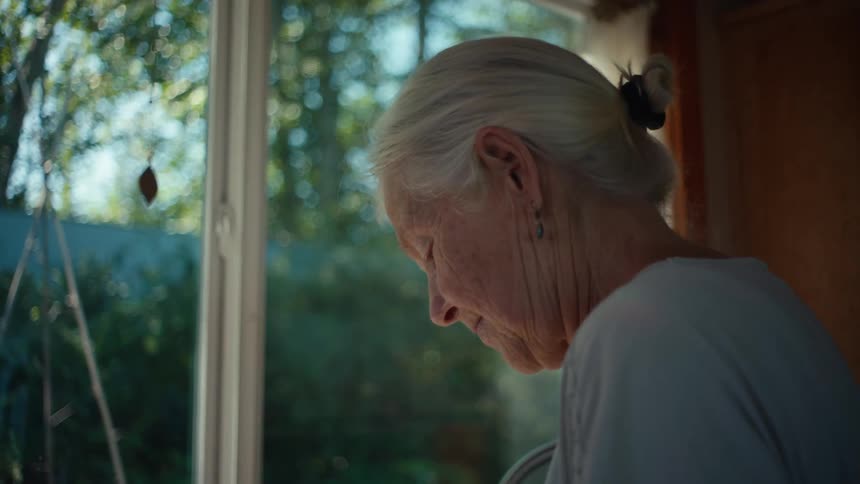 The face of an elderly woman with gray hair tied in a ponytail and she is looking down while fiddling with something with her hands