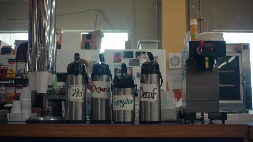 Four silver thermoses labeled dark regular organic and decaf sit on a counter next to a coffee machine