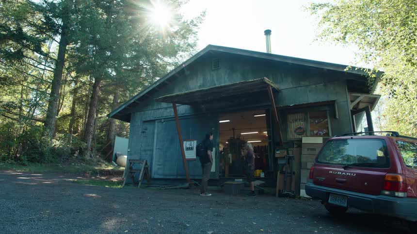 A rustic workshop surrounded by trees with sunlight shining through the branches a red subaru parked nearby and two people talking at the entrance