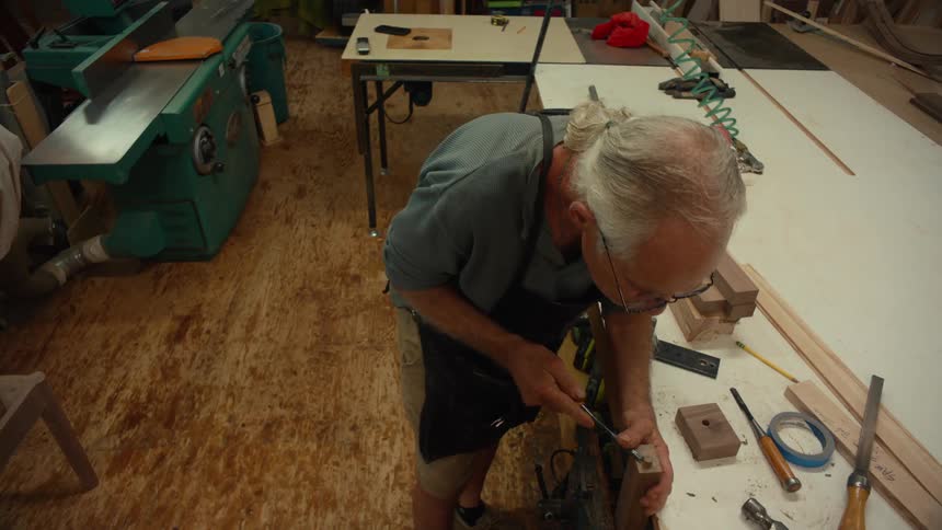 A man with glasses and tied back gray hair working in a woodworking shop surrounded by tools wooden pieces and machinery on a wellworn floor