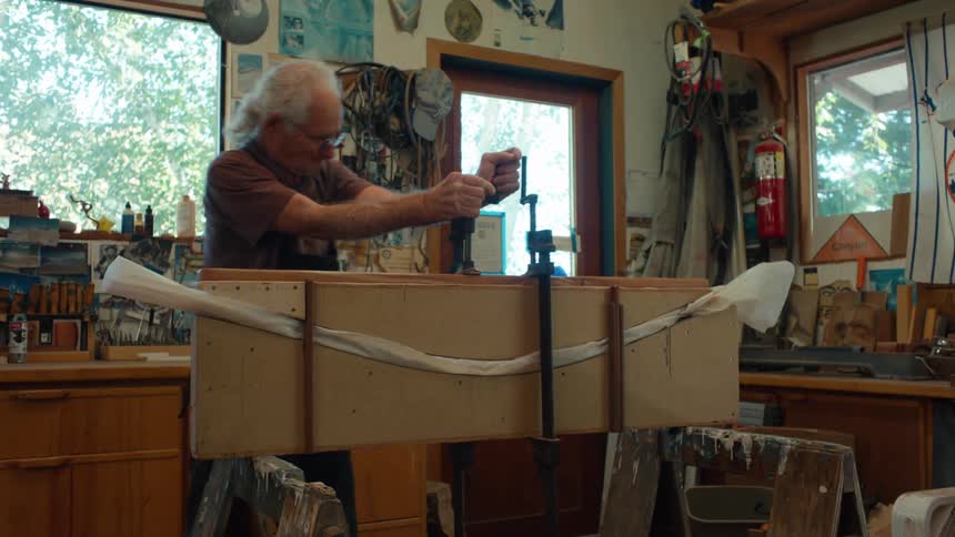 An elderly craftsman working in a workshop using a clamp on a wooden project surrounded by tools and a creative workspace