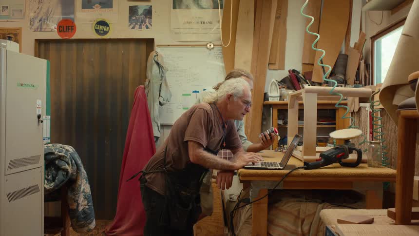 Two men at a workbench in a workshop consult a container while using a laptop