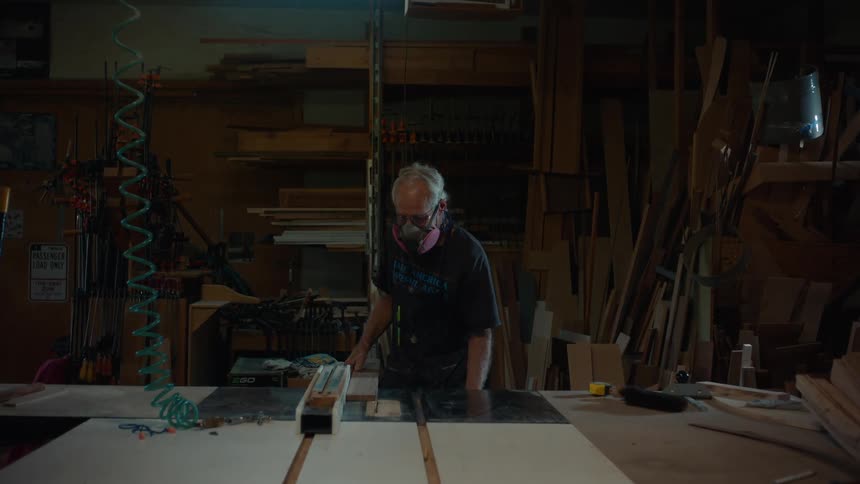 An elderly carpenter man is wearing a protective mask while working on a wooden product that is in front of him