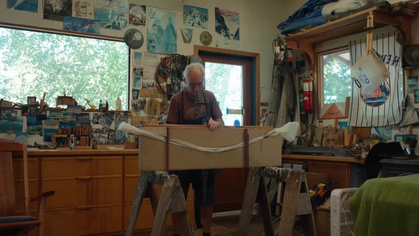 An elderly carpenter positions wood on saw horses and then applies clamps to hold the pieces into place