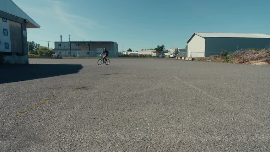 Two cyclists ride across a wide empty paved lot under a clear blue sky with industrial buildings in the background
