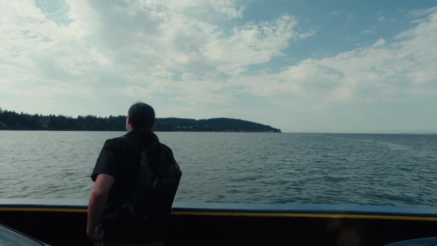 A person with a backpack standing on a ferry or boat looking out over a calm body of water toward a distant treecovered shoreline under a partly cloudy sky