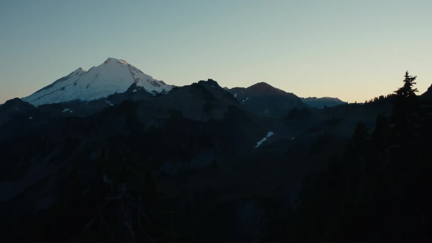 A serene mountain peak capped with snow rises against a twilight sky surrounded by dark rugged hills