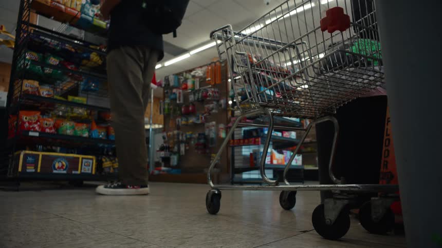 A shopping cart and a customer are seen in a brightly lit convenience store stocked with various goods