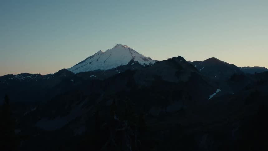 A snow capped mountain stands tall against the twilight sky towering over the surrounding peaks