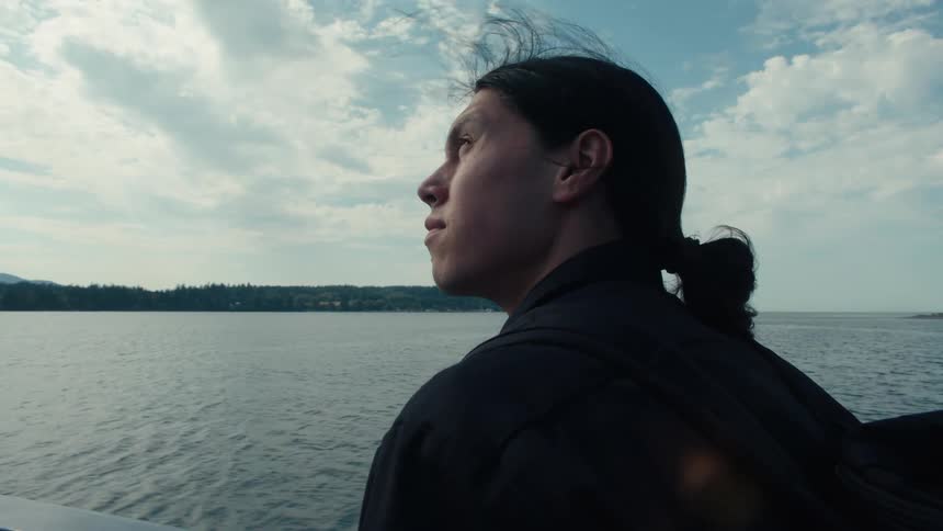 A man with long hair and a black coat is seated on a boat looking contemplatively towards the expansive ocean ahead