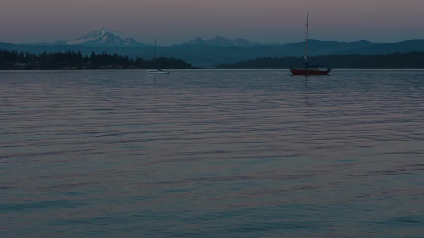 Two sailboats are anchored in a calm bay at dusk with a snowcapped mountain range visible in the distance