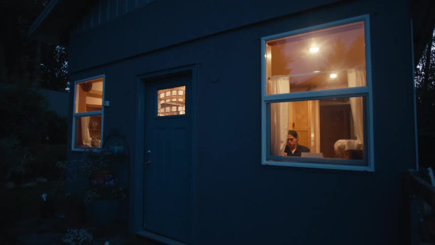 A woman and an elderly person are shown sitting in their kitchen being viewed through a window outside