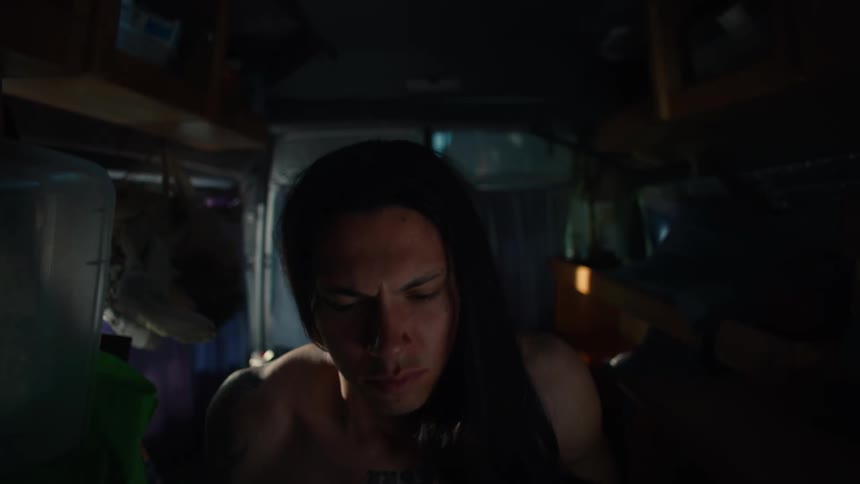 A young shirtless man with long hair wakes up inside a camper