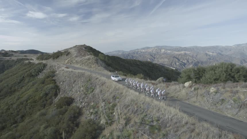 A car following a bunch of bicyclists on a country road around mountains