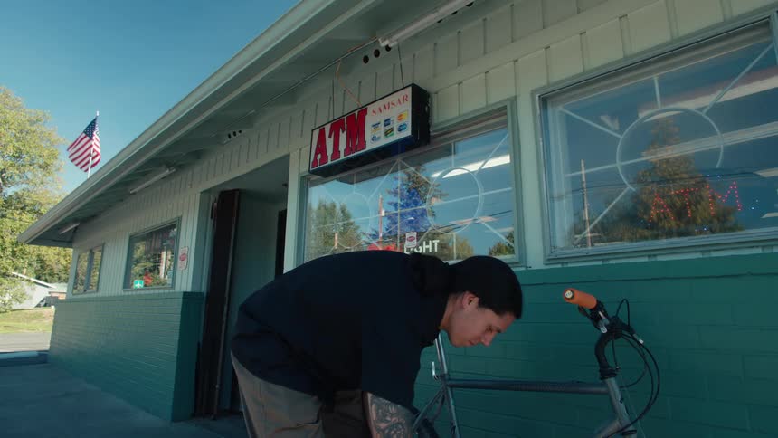 A young man in a black shirt leaves a store with some objects in his hand and puts them in his backpack and then takes his bicycle that is leaning against the wall and leaves